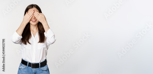 Smiling happy woman waiting for surprise, shut eyes with hands, standing blindsided against white background photo