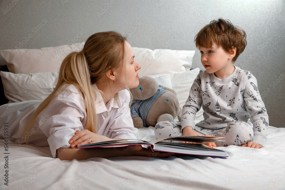 Cute preschool child son holding a book, reading fairy tales for mom, lying in bed together, happy mother listening to a focused little boy who is learning to read, enjoy bedtime stories Cute preschool child son holding a book, reading fairy tales for mom, lying in bed together, happy mother listening to a focused little boy who is learning to read, enjoy bedtime stories