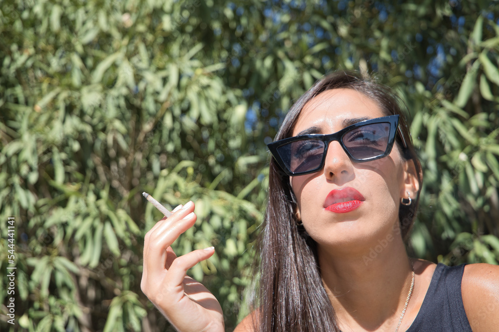 Young woman, brunette, slender, dressed in black and wearing sunglasses, sitting on a bench exhaling cigarette smoke while smoking. Concept tobacco, cigarette, addiction, health, smoke. Young woman, brunette, slender, dressed in black and wearing sunglasses, sitting on a bench exhaling cigarette smoke while smoking. Concept tobacco, cigarette, addiction, health, smoke.