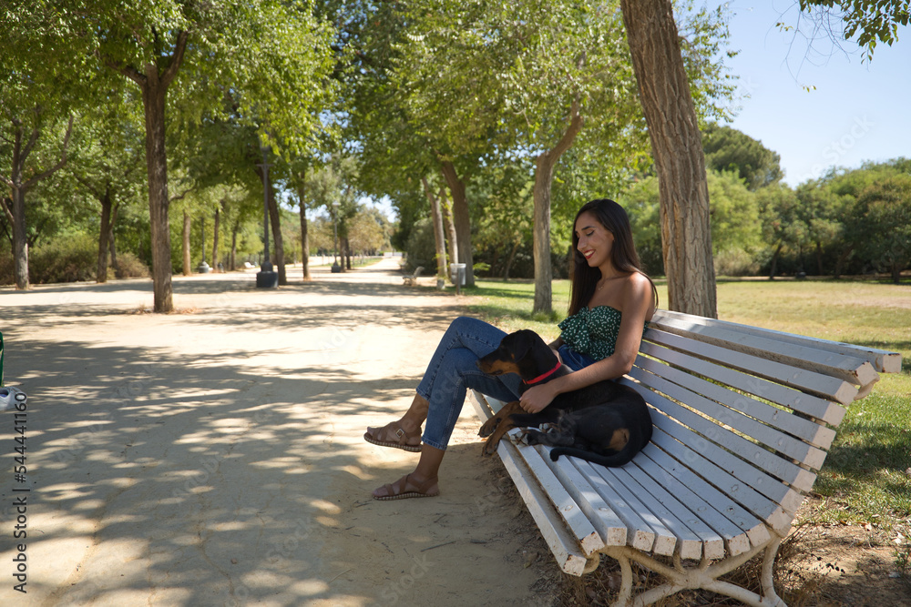 Young woman, brunette, slender, dressed in green t-shirt and jeans, sitting on a bench next to her dog, happy and in a loving attitude. Concept animals, pets, friends, love, dogs, breed.