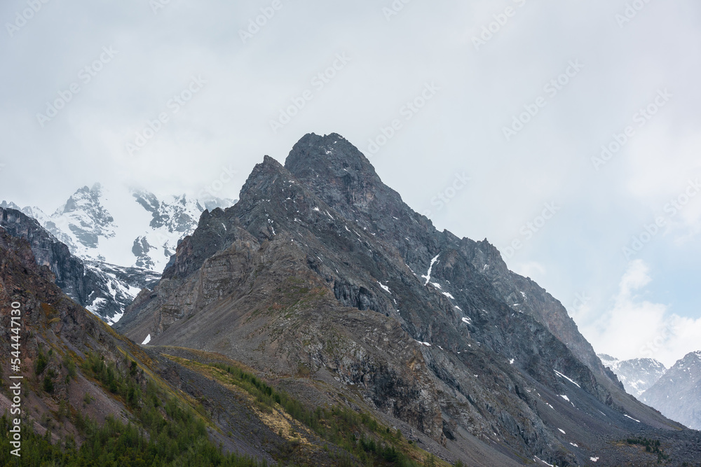 Atmospheric mountain landscape with large pointy pinnacle under gray cloudy sky. Gloomy scenery with high pointed mountain peak with sharp rocks in low clouds. Huge rocky peaked top in rainy weather.