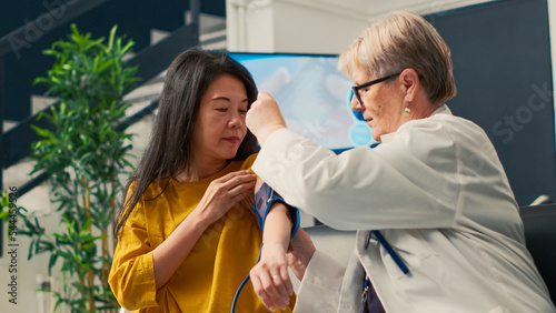 Elderly medic measuring hypertension and pulse pressure with tonometer on asian old woman in waiting room. Using cardiology instrument to check hypotension and cardiac exam. Handheld shot. photo