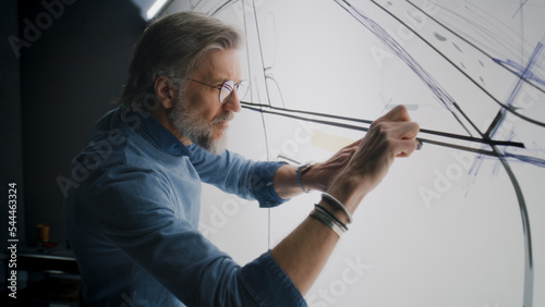 Automotive male engineer creating sketch with duct tape on paper whiteboard, developing futuristic new generation electric car. He working in modern car design development studio.