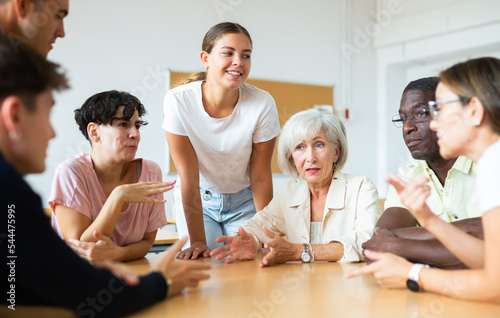 Group of positive people of different ages and nationalities sitting around table and communicating in English-speaking club with young female tutor..