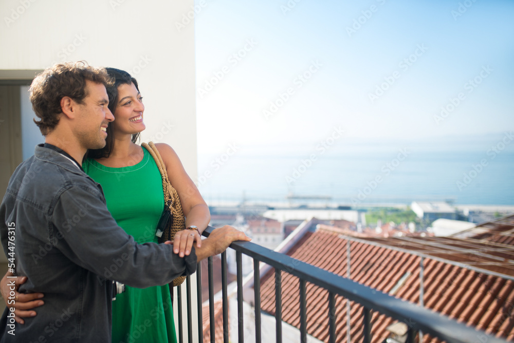 Side view of young multiethnic couple resting on balcony. Handsome man and beautiful woman standing close embracing and enjoying faraway city views from height. Romance, love, summer vacation concept