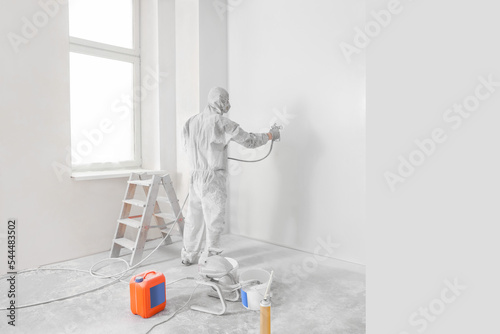 Decorator in uniform painting wall with sprayer indoors