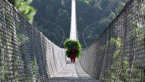 A Nepali person carrying a heavy load and walking across a suspension bridge over a valley in Nepal.