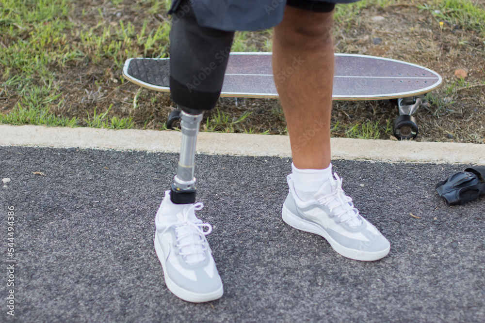Close-up of prosthetic leg in sneakers. White shoes and skateboard, man training in park. Sport, disability concept