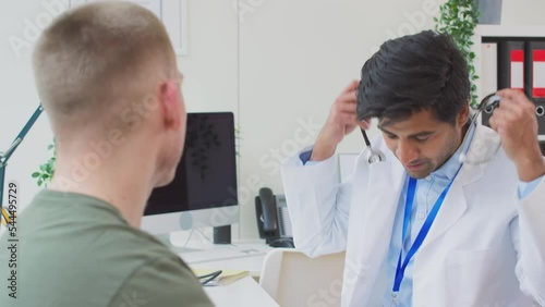 Male doctor or GP wearing white coat examining young man listening to heartbeat with stethoscope- shot in slow motion photo