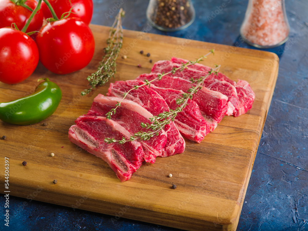 Creative culinary composition. Appetizing meat steaks with a sprig of fragrant rosemary on a cutting board, spices, seasonings, ripe tomatoes and peppers on a dark blue background. Close-up. Creative culinary composition. Appetizing meat steaks with a sprig of fragrant rosemary on a cutting board, spices, seasonings, ripe tomatoes and peppers on a dark blue background. Close-up.