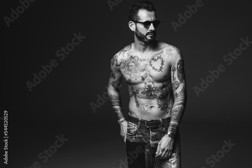 Portrait of handsome confident stylish hipster lambersexual model. Sexy modern man. Naked torso with tattoos.Fashion male posing in studio on dark background in sunglasses © halayalex