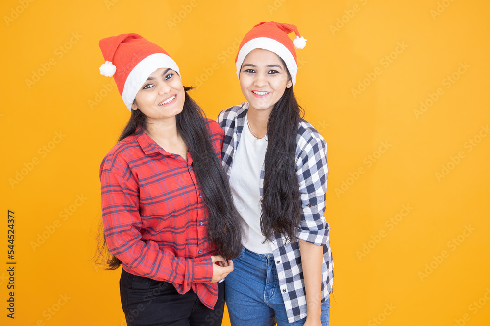 Happy indian girls wearing christmas hat laughing together isolated yellow orange background. X-mas celebration. Winter season.