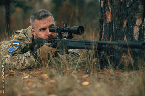 Ukrainian soldier dressed military uniform lying on ground with sniper rifle with silencer at front line. Handsome brave confident man at war during russian military invasion of Ukraine. Closeup.