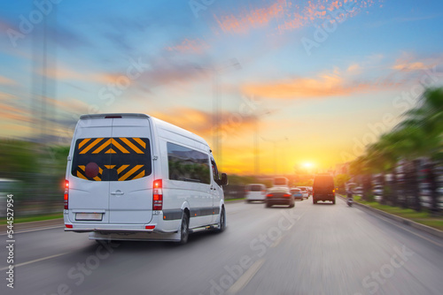 Unrecognizable white small passenger van hurry up on highway at city street traffic with urban cityscape, sunrise sky on background. Charter fast bus van hurry up on with motion blur effect