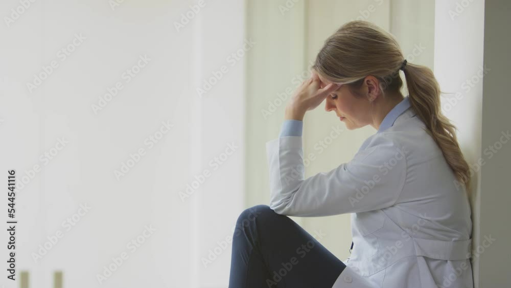Stressed female doctor wearing white coat sitting in hospital corridor with head in hands - shot in slow motion Stressed female doctor wearing white coat sitting in hospital corridor with head in hands - shot in slow motion