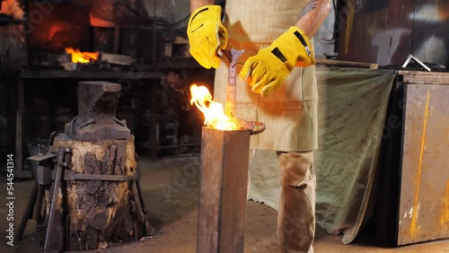 A male blacksmith dips a red-hot sword into a container with oil for quenching metal. The process of working in a blacksmith shop.