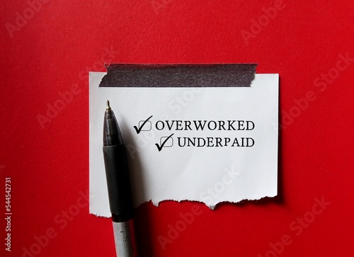 Torn note on red background with handwritten text OVERWORKED and UNDERPAID, means employees or workers work too hard or too long hours but get paid less than what they deserve photo