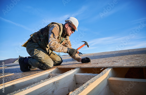 Carpenter hammering nail into OSB panel on the roof top of future cottage on blue sky background. Man worker building wooden frame house. Carpentry and construction concept.
