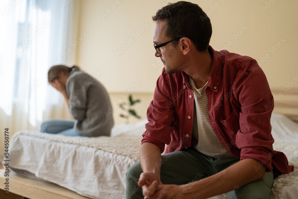 Confused man looks towards crying girl sits on opposite side of bed after unpleasant conversation. Young married couple needs help of professional psychologist because quarrels and misunderstandings Confused man looks towards crying girl sits on opposite side of bed after unpleasant conversation. Young married couple needs help of professional psychologist because quarrels and misunderstandings