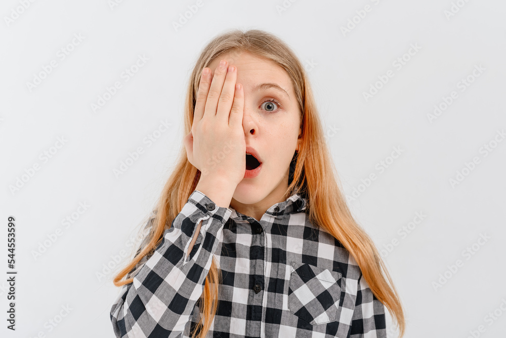 shocked and concerned teen girl open mouth and covers half of face with palm, looks with one eye at camera, stands over white studio background shocked and concerned teen girl open mouth and covers half of face with palm, looks with one eye at camera, stands over white studio background