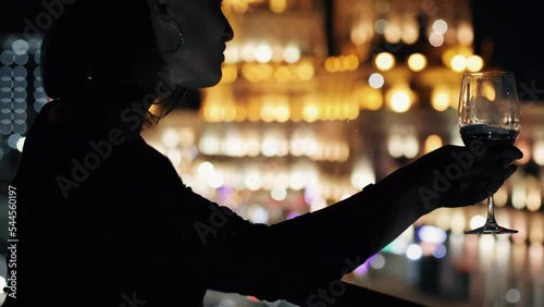 Elegant brunette woman drinking white wine glass at rooftop bar terrace looking at city lights. Sexy female hanging out. Big city night concept.