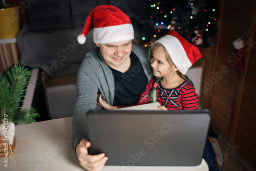 Safe online Christmas celebration. Happy family in Santa red hats celebrating with friends virtually via internet and notebook. Video call. Stay home, distant holiday, lifestyle