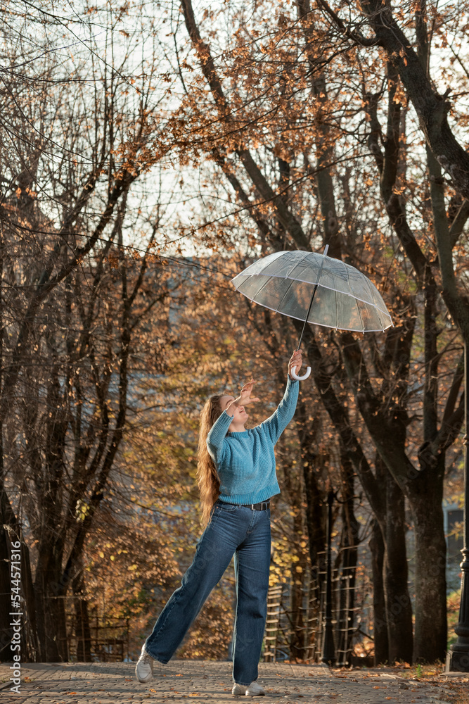 Cheerful girl in blue sweater and pants with transparent umbrella on alley. Beautiful woman dance in autumn park Cheerful girl in blue sweater and pants with transparent umbrella on alley. Beautiful woman dance in autumn park
