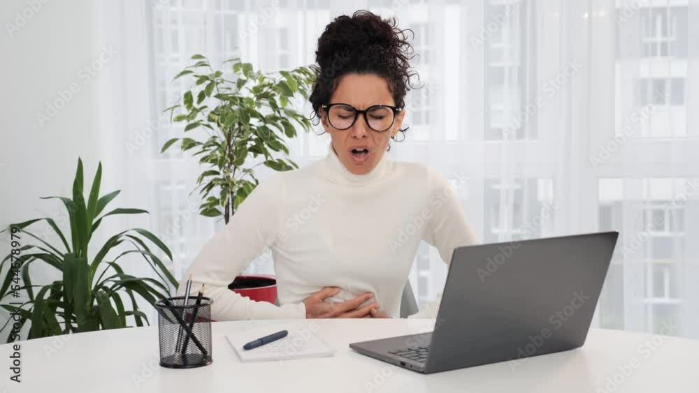 Focused latin woman in glasses wearing white sweater working on laptop while clasping belly suffering from menstrual, severe abdominal pain, periods spasm, irritable bowel syndrome of diet at home.