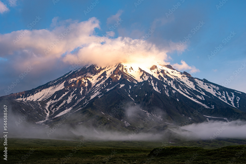 Smoking volcano covered with snow, green valley, blue sky with clouds, Russia, Kamchatka. The sun illuminates the volcano.