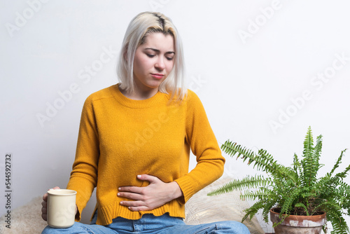 Blonde woman sitting on ground holding cup of tea and one hand on belly