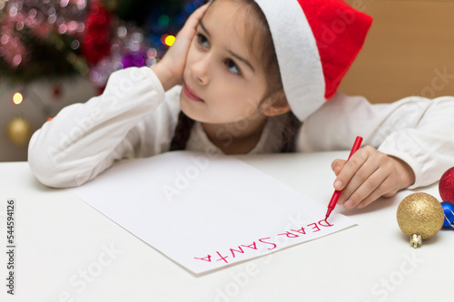 Cute little girl in Santa hat is writing a letter to Santa Claus on Christmas. Shallow depth of field, focus on the hand