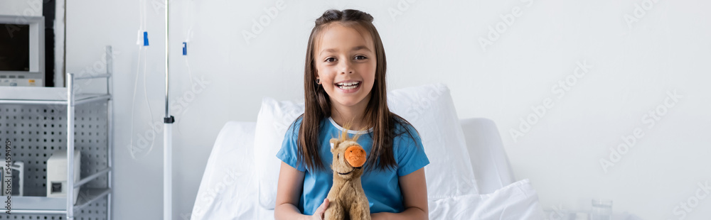 Cheerful kid holding soft toy and looking at camera in clinic, banner. Cheerful kid holding soft toy and looking at camera in clinic, banner.