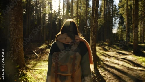 Camera follows from behind a young woman walking in the beautiful forest, exploring beautiful nature. Wearing fleece and backpack. Sun is shinning in the camera, lens flare. photo