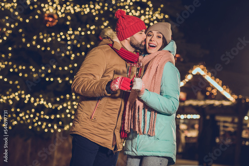 Portrait of two idyllic partners hold champagne kiss cheek enjoy x-mas evening advent lights outside