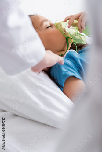 Blurred pediatrician touching child with oxygen mask on bed in clinic.