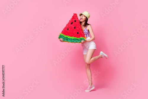 Full size photo of adorable cheery young lady hold bite watermelon pinata wear stylish tank top headwear isolated on pink color background