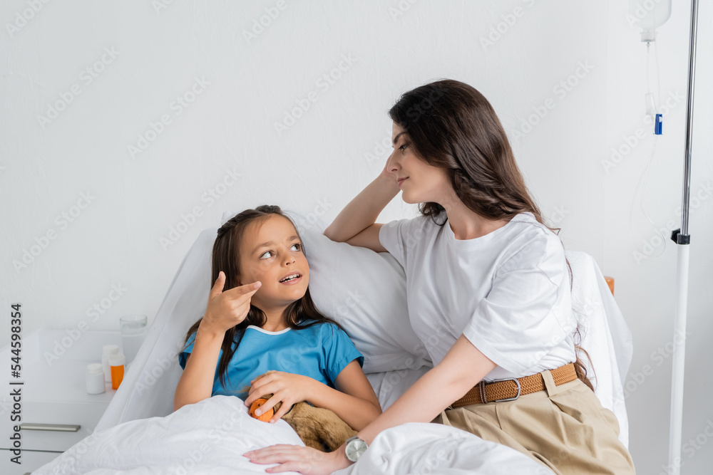 Fototapeta premium Kid with soft toy talking to mother on bed in hospital.