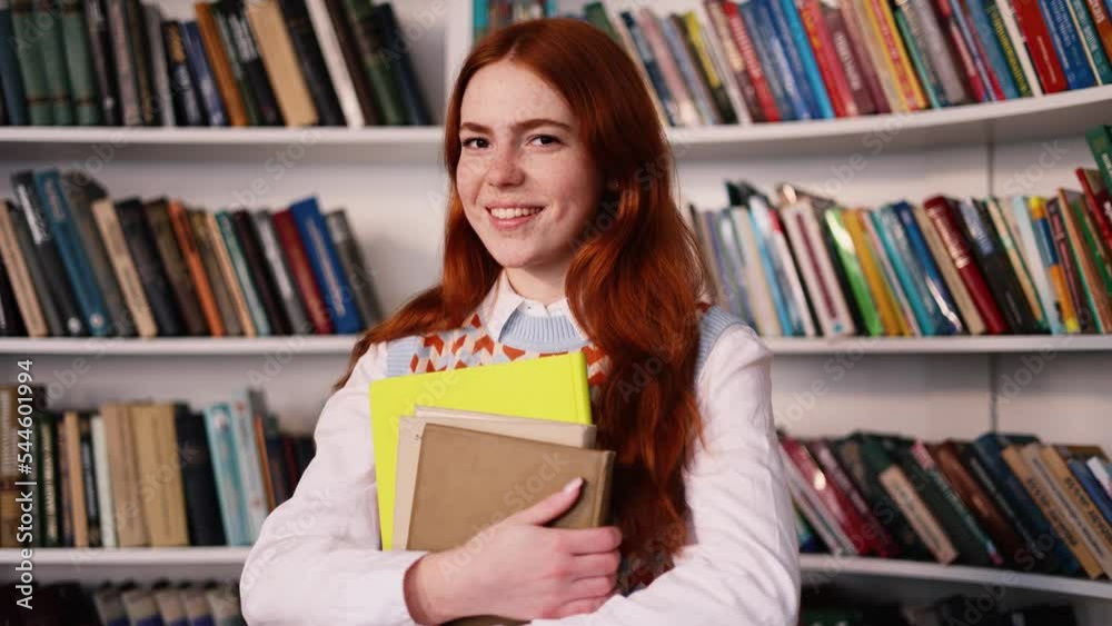 Portrait of charming red hair girl with freckles looking at the camera at the university library. Young student with book on her hands preparing for exams in the library. Portrait of charming red hair girl with freckles looking at the camera at the university library. Young student with book on her hands preparing for exams in the library.