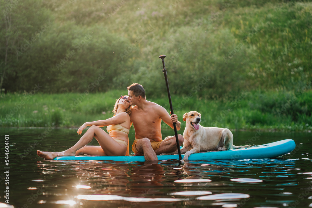 Young couple in love sitting on pad board with dog on river at summertime during vacations kissing. Weekend and outdoor leisure activities. People and domestic animals. Husband kissing wife. Young couple in love sitting on pad board with dog on river at summertime during vacations kissing. Weekend and outdoor leisure activities. People and domestic animals. Husband kissing wife.