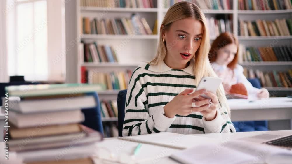 Attractive young student look at the phone screen and show thumbs up at university library. Girl looking approvingly at camera and recommend it. Attractive young student look at the phone screen and show thumbs up at university library. Girl looking approvingly at camera and recommend it.