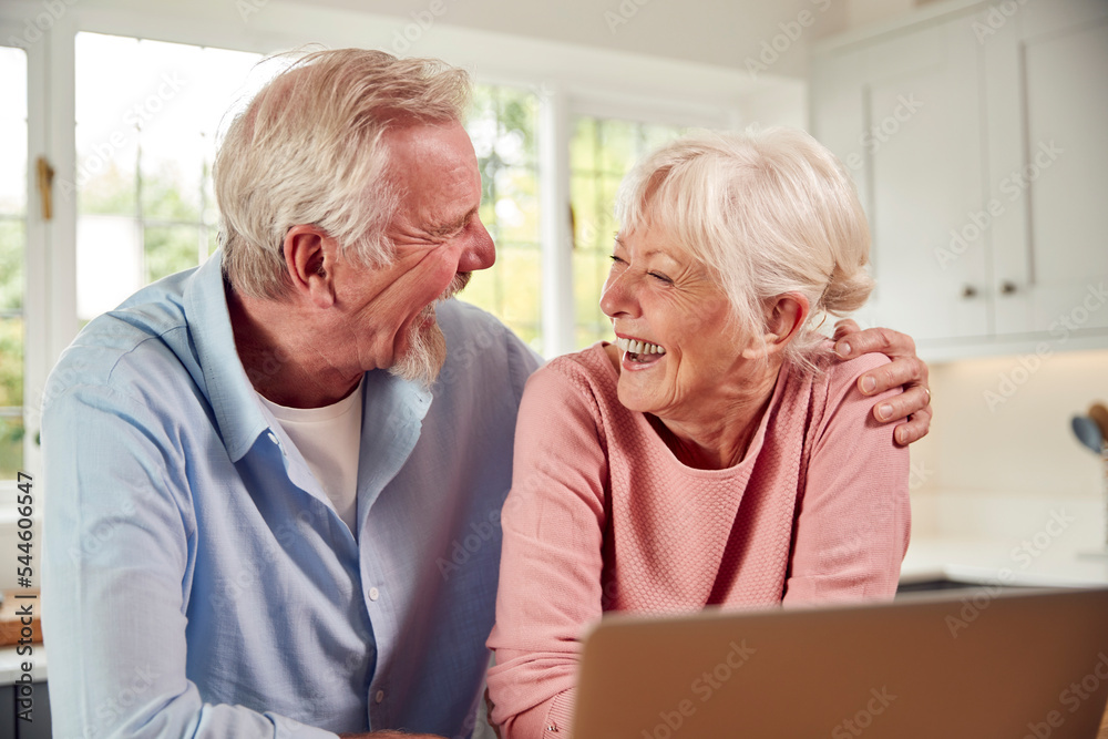Retired Senior Couple Sitting In Kitchen At Home Using Laptop