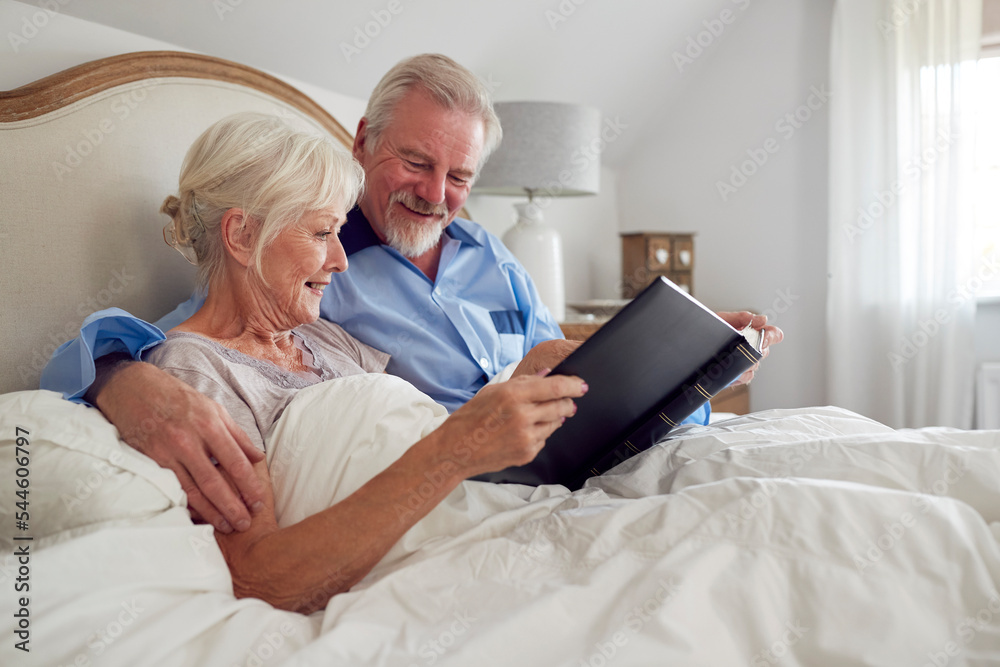 Retired Senior Couple In Bed At Home Looking At Photo Album Together