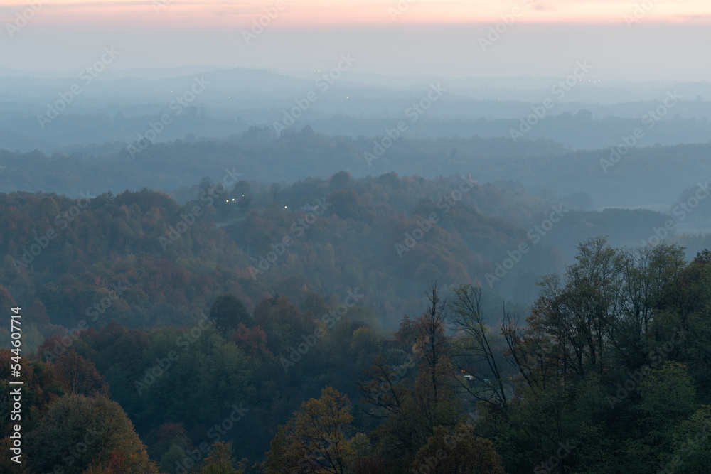 Hilly village with lush forest during foggy evening in autumn, rural landscape with hill layers in distance Hilly village with lush forest during foggy evening in autumn, rural landscape with hill layers in distance
