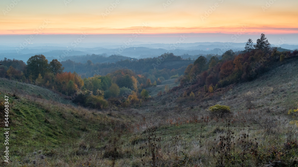 Rural landscape during dusk in autumn, hilly countryside with lush colourful forests and orange glow in sky