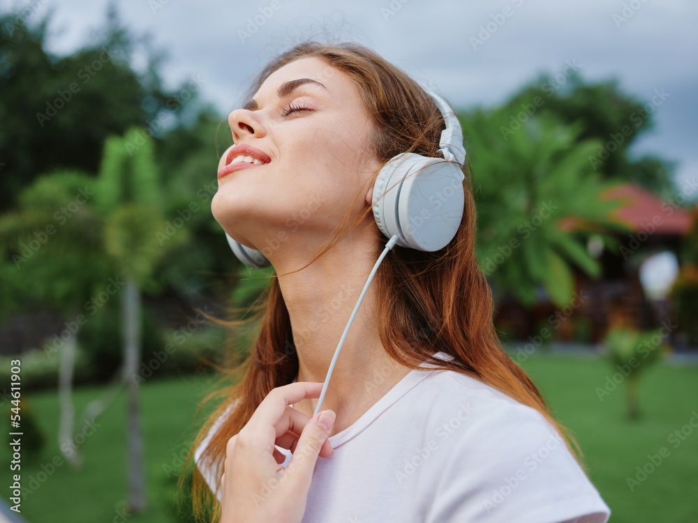 Woman portrait in headphones happiness smile with her eyes closed in a white T-shirt listening to music and walking down the street, in front of palm trees, meditation and tranquility Woman portrait in headphones happiness smile with her eyes closed in a white T-shirt listening to music and walking down the street, in front of palm trees, meditation and tranquility