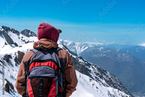 rear view of young man hiker with backpack looking at view of snowy peaks of mountains in winter against blue sky hiking copy space enjoying nature