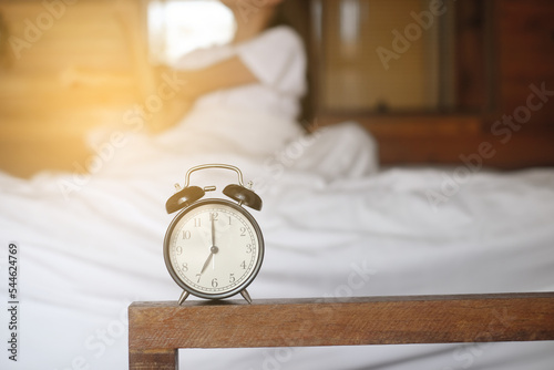 Fototapeta Closeup alarm clock having a good day with background happy woman stretching in bed after waking up, sunlight in morning
