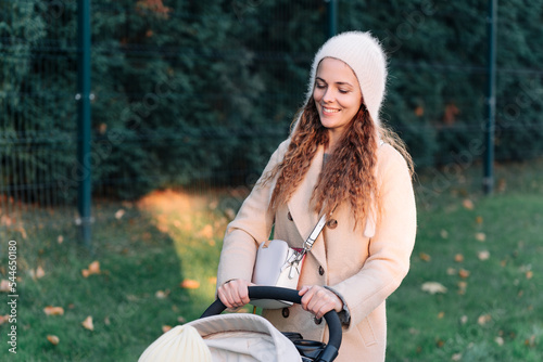 Young woman in a beige coat walks with a child in a stroller through the town. Mother with pram. Woman with baby stroller walks in the park at sunset.