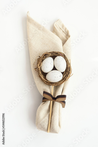 Decorative wicker nest with eggs and folded napkin on white background upper view. Festive table setting for Easter holiday celebration