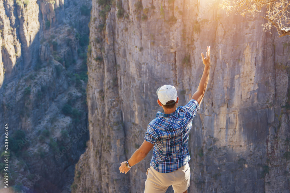 Man in checkered blue shirt and cap stands on the edge of deep canyon cliff, raising one hand up, looking down in surprise at the beautiful landscape around.